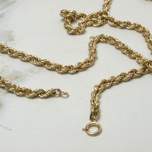 May include: A gold-colored rope chain necklace with a spring ring clasp. The chain is made of twisted links, creating a rope-like appearance. The necklace is displayed on a light-colored surface.