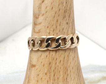 May include: A gold-toned ring featuring a continuous curb chain link design. The metal band has a polished, reflective surface and is displayed on a light-colored wooden cone stand. The ring consists of interlocking oval links that form a uniform band.