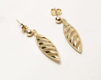May include: A pair of gold-colored earrings. Each earring features a post and a small ball, with a long, leaf-shaped pendant. The pendant has a series of parallel, curved cutouts, creating an open design. The earrings are set against a white background.