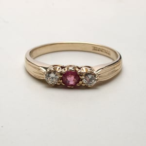 May include: A gold ring with three stones. The center stone is a pink ruby, and the two stones on either side are diamonds.