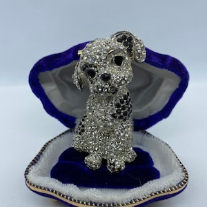 Puede incluir: Una figura de cachorro enjoyada con cristales negros y transparentes se encuentra dentro de un joyero en forma de concha de terciopelo azul. El cachorro tiene manchas y ojos negros. La caja tiene un borde dorado.