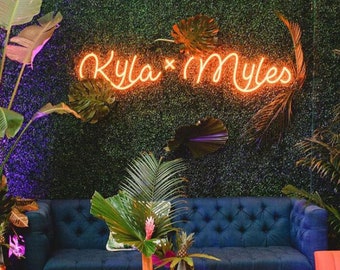May include: A blue sofa with a neon sign that reads "Kyla x Myles" above it. The sofa is in front of a green wall with tropical plants. There is a wooden coffee table in front of the sofa and a pink chair in the foreground.