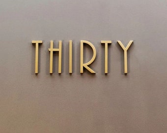 Könnte beinhalten: Ein goldfarbenes Metallschild mit dem Wort "THIRTY" in einer stilisierten Schrift.