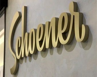Könnte beinhalten: Goldfarbenes, dreidimensionales Schild mit dem Wort "Schoener" in Schreibschrift. Das Schild ist an einer strukturierten, hellgrauen Wand befestigt. Die Buchstaben werfen einen Schatten.