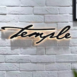 May include: A black cursive sign with the word "Temple" illuminated with white light against a white brick wall.