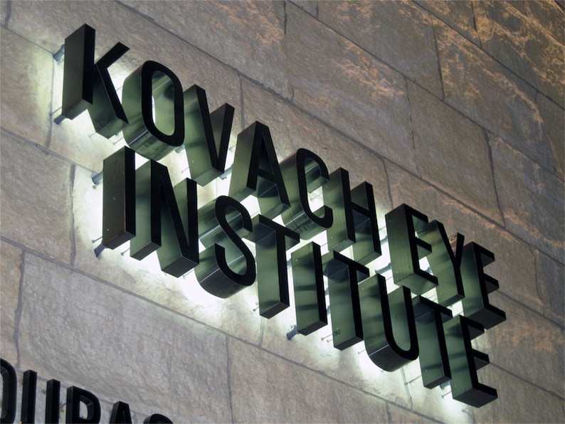 以下が含まれることがあります： 文字の後ろに白いライトが付いた黒い看板に「Kovachey Eye Institute」と書いてあります。