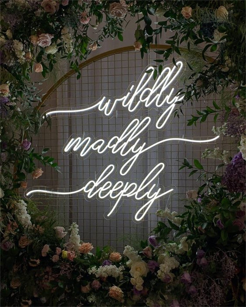 May include: White neon sign with the words "wildly madly deeply" in a cursive font. The sign is hanging in front of a gold wire mesh backdrop decorated with flowers.