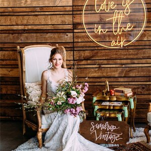 May include: A bride in a white lace wedding dress sits in a vintage chair with a bouquet of flowers. A neon sign that reads "Love is in the air" hangs on a wooden wall behind her. A wooden sign with the text "Sundrop Vintage" is on the floor in front of her.