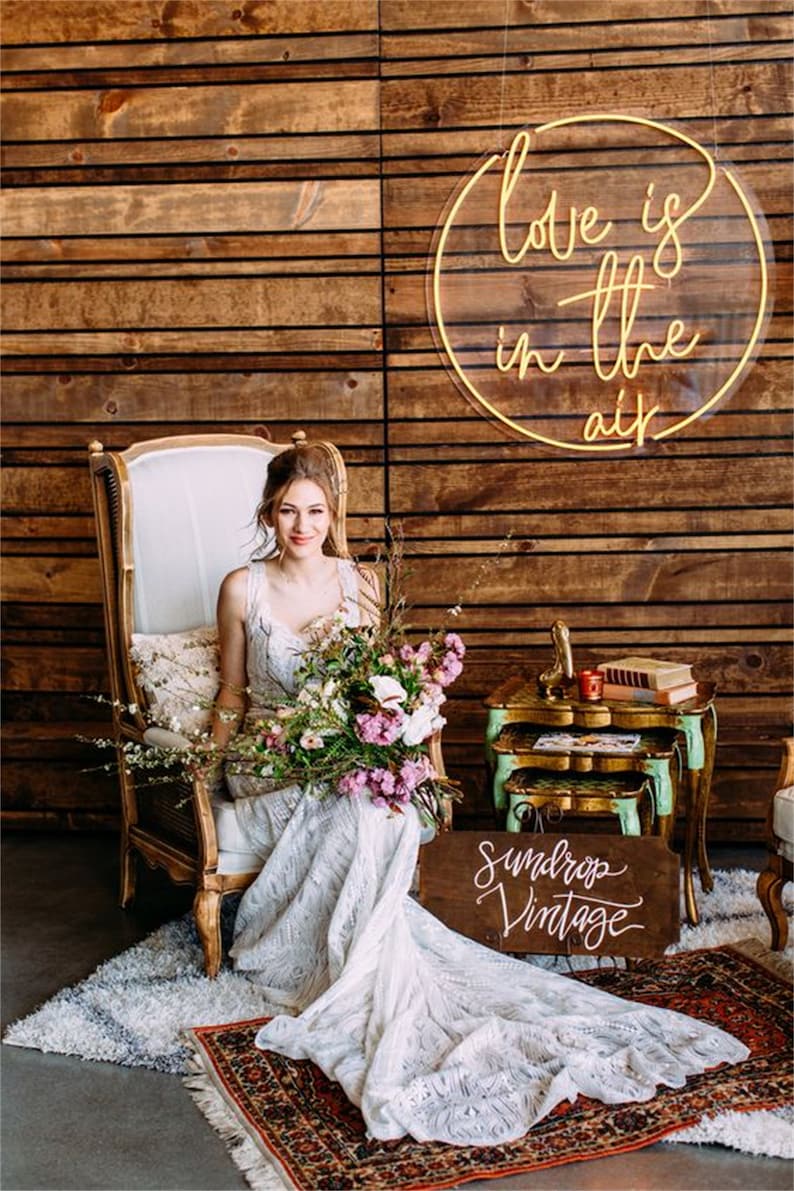 May include: A bride in a white lace wedding dress sits in a vintage chair with a bouquet of flowers. A neon sign that reads "Love is in the air" hangs on a wooden wall behind her. A wooden sign with the text "Sundrop Vintage" is on the floor in front of her.