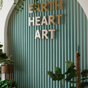 K&ouml;nnte beinhalten: Eine tealgr&uuml;ne Wand mit vertikalen Rillen, auf der ein goldenes Schild mit der Aufschrift "EARTH HEART ART" angebracht ist. Die Wand ist mit Topfpflanzen und Gr&uuml;npflanzen dekoriert.