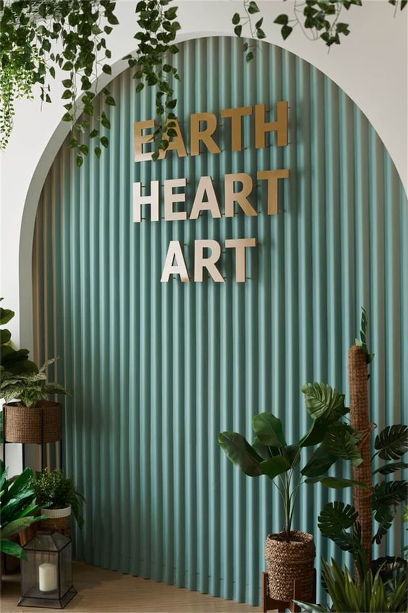 K&ouml;nnte beinhalten: Eine tealgr&uuml;ne Wand mit vertikalen Rillen, auf der ein goldenes Schild mit der Aufschrift "EARTH HEART ART" angebracht ist. Die Wand ist mit Topfpflanzen und Gr&uuml;npflanzen dekoriert.