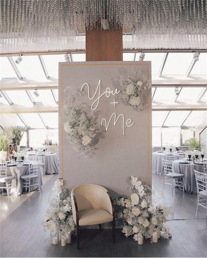 May include: A wedding backdrop with a white neon sign that says "You + Me" surrounded by white flowers. A white chair sits in front of the backdrop.