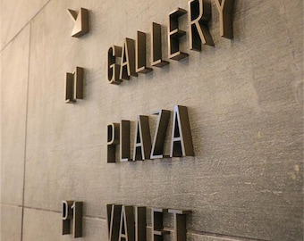 Puede incluir: Un letrero con letras doradas en una pared gris. El letrero dice "Gallery", "Plaza" y "P1 Valet".