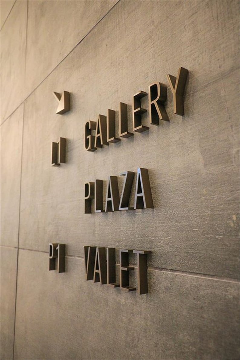 K&ouml;nnte beinhalten: Ein Schild mit goldenen Buchstaben an einer grauen Wand. Das Schild zeigt "Gallery", "Plaza" und "P1 Valet".