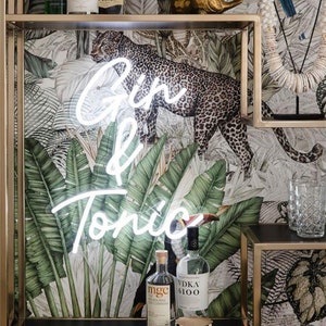 May include: A white neon sign that says "Gin & Tonic" is hanging on a wall with a tropical wallpaper featuring a leopard and toucans. There are two bottles of liquor on a shelf below the sign.