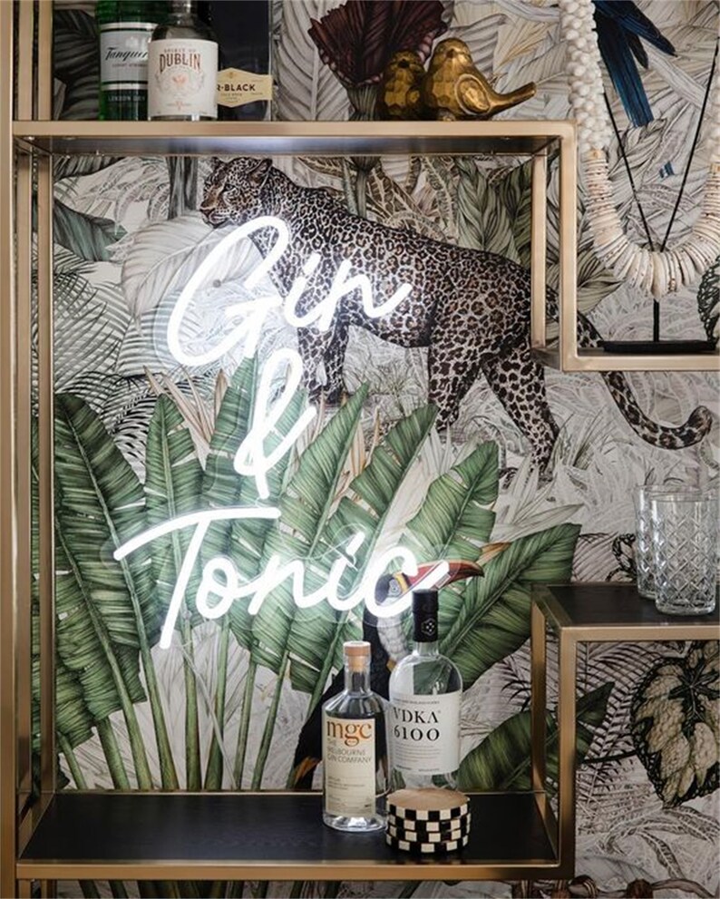 May include: A white neon sign that says "Gin & Tonic" is hanging on a wall with a tropical wallpaper featuring a leopard and toucans. There are two bottles of liquor on a shelf below the sign.