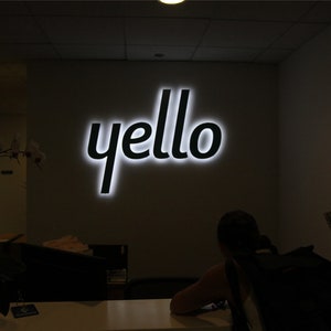 以下が含まれることがあります： 白黒の看板に、白い輪郭で「yello」という文字が書かれている。看板は点灯しており、暗い部屋の壁に取り付けられている。