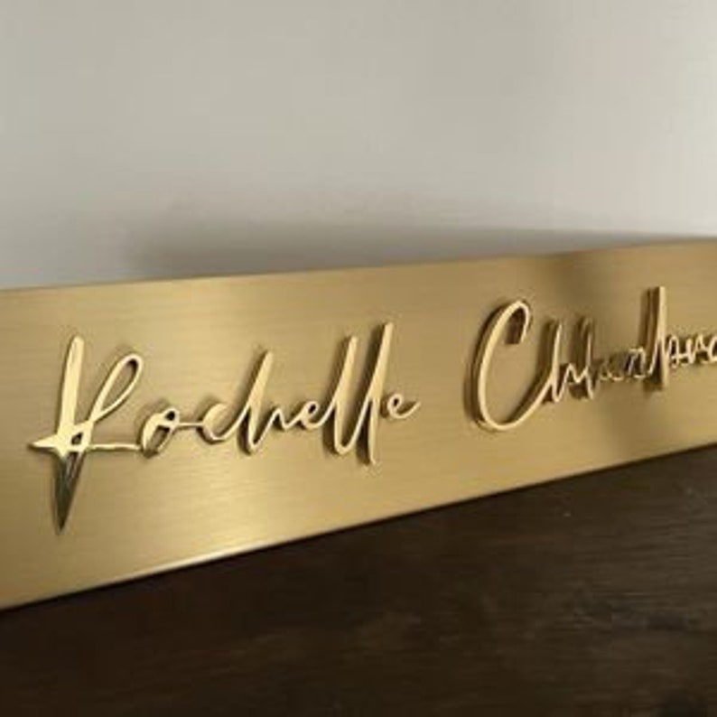 May include: A gold-coloured rectangular sign with the name "Rochelle Christopher" in a cursive font. The sign is made of a brushed metal material and is displayed on a dark wooden surface.