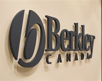 Könnte beinhalten: Ein dunkelgraues 3D-Schild an einer beigefarbenen Wand. Das Schild trägt die Aufschrift "Berkley CANADA" in einer Serifenschrift. Das "B" in "Berkley" ist eine stilisierte ovale Form. Die Buchstaben sind erhaben und werfen einen Schatten.
