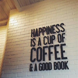 May include: A white brick wall displays the quote "HAPPINESS IS A CUP OF COFFEE & A GOOD BOOK" in black, bold lettering. The wall is illuminated with warm lighting, and the ceiling is constructed from wooden planks.