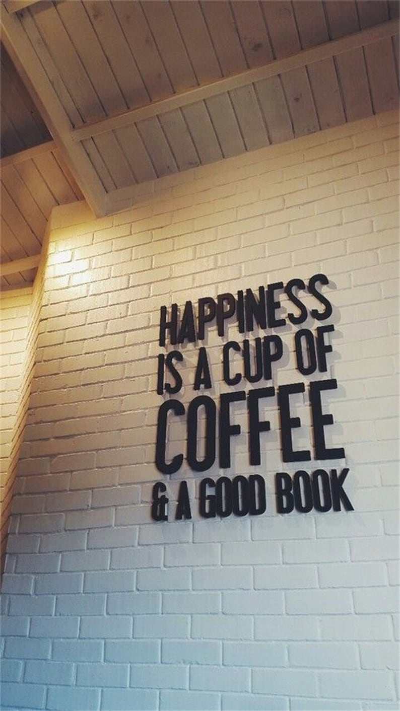 May include: A white brick wall displays the quote "HAPPINESS IS A CUP OF COFFEE & A GOOD BOOK" in black, bold lettering. The wall is illuminated with warm lighting, and the ceiling is constructed from wooden planks.