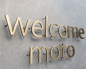Puede incluir: Letras de metal de color dorado que deletrean "Welcome moto" montadas en una pared gris.