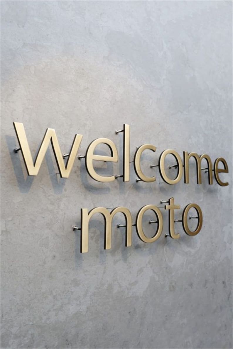 May include: Gold-coloured metal letters spell "Welcome moto" on a grey concrete wall. The letters are mounted with small silver pins, creating a modern, minimalist design. The word "Welcome" is above "moto".