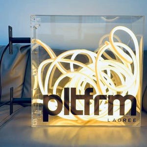 Pode incluir: Uma caixa de acrílico transparente com uma luz de néon branca dentro. A caixa tem um logotipo preto que diz "pltfrm LAGREE".