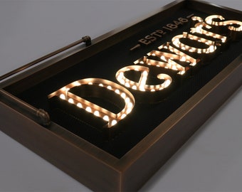 May include: A vintage-style sign with the text "ESTD 1846" and the word "Dewar's" in illuminated letters. The sign is made of metal and has a dark brown finish.