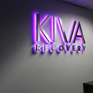 以下が含まれることがあります： モダンなフォントで「KIVA RECOVERY」と書かれた銀色の看板。文字は紫色のネオンで縁取られています。