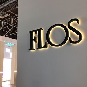 以下が含まれることがあります： 白い壁に「FLOS」と書かれた黒い照明付きの看板があります。