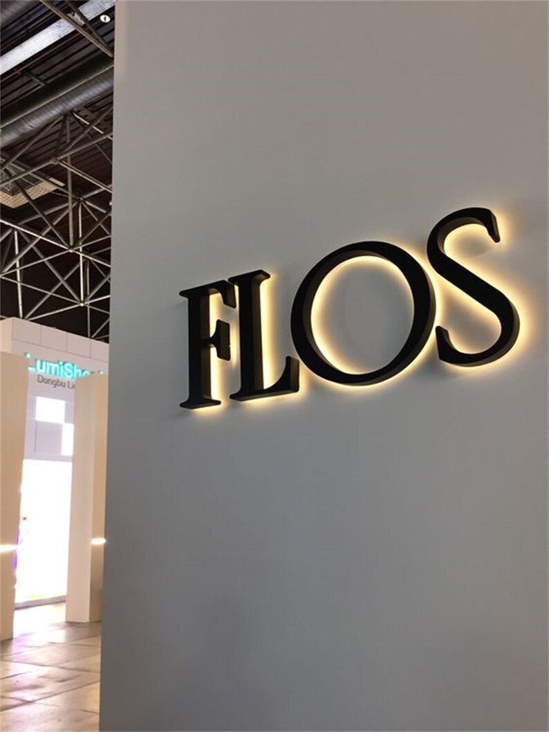 以下が含まれることがあります： 白い壁に「FLOS」と書かれた黒い照明付きの看板があります。