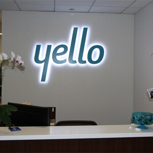 以下が含まれることがあります： 白い受付カウンターの後ろの壁に、ターコイズブルーの「yello」ロゴがあります。ロゴは白いライトで照らされています。