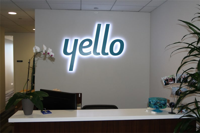 以下が含まれることがあります： 白い受付カウンターの後ろの壁に、ターコイズブルーの「yello」ロゴがあります。ロゴは白いライトで照らされています。