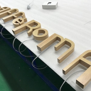 Könnte beinhalten: Holzbuchstaben, die das Wort "Coffee Roma" in einer hellbraunen Farbe buchstabieren. Die Buchstaben sind von hinten beleuchtet und erzeugen ein warmes Leuchten.