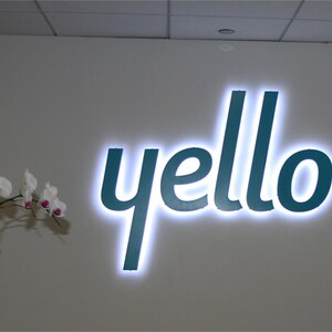 以下が含まれることがあります： 白い壁に、白く縁取られたターコイズブルーの「yello」の看板が取り付けられています。