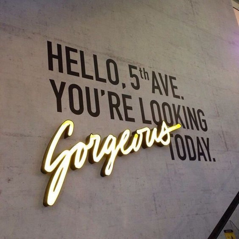 以下が含まれることがあります： 白い壁に「Hello, 5th Ave. You're looking gorgeous today.」という黒い文字が書かれている。テキストの下には、「gorgeous」という黄色のネオンサインがある。