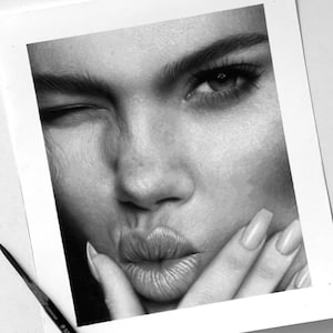 Puede incluir: Un detallado dibujo a lápiz en blanco y negro del rostro de una persona, guiñando un ojo y con los labios fruncidos. El dibujo está sobre papel blanco, con un lápiz visible en la esquina inferior izquierda. La textura de la piel y los rasgos faciales están meticulosamente representados.