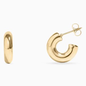 May include: A pair of gold-colored, chunky hoop earrings. One earring is shown upright, and the other is on its side, displaying the post and clasp. The earrings have a smooth, polished finish and a rounded, tubular design.
