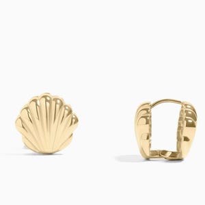 May include: A pair of gold-colored seashell-shaped earrings. One earring is a stud, and the other is a hinged hoop. The earrings have a polished finish and are set against a white background. The earrings are a jewelry item.