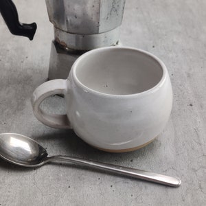 May include: A white ceramic mug with a rounded shape and a handle. A silver spoon is lying on a grey surface next to the mug.