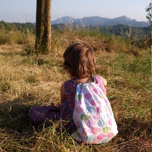 May include: A young person sits in a field of long grass, wearing a purple dress and a pink and white drawstring backpack with a polka dot pattern. The person is looking out at a distant mountain range.
