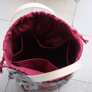 May include: A burgundy fabric bag with a drawstring closure and a cream-colored leather handle. The bag has three compartments inside and a floral print on the exterior.