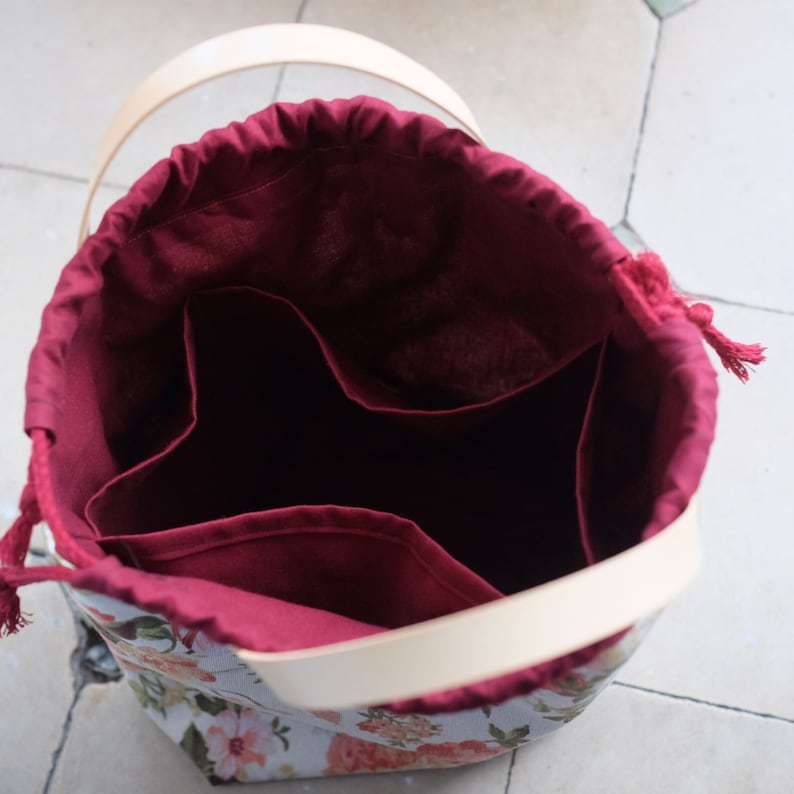 May include: A burgundy fabric bag with a drawstring closure and a cream-colored leather handle. The bag has three compartments inside and a floral print on the exterior.
