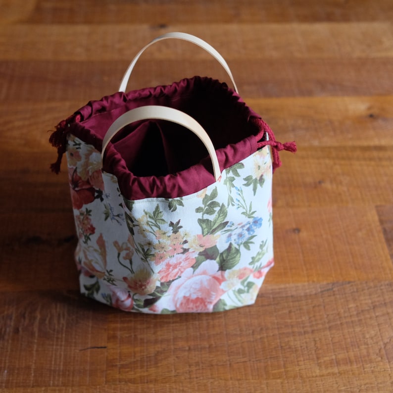 May include: A white fabric bag with a floral print and a burgundy lining. The bag has two wooden handles and a drawstring closure.