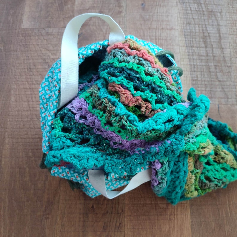 May include: A teal, green, and pink crocheted bag with a white fabric handle. The bag is made of a soft, textured material and has a floral pattern.