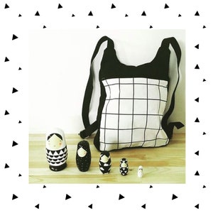 May include: A black and white checkered fabric backpack with a black flap and adjustable straps. Five black and white nesting dolls are in front of the backpack on a wooden surface.