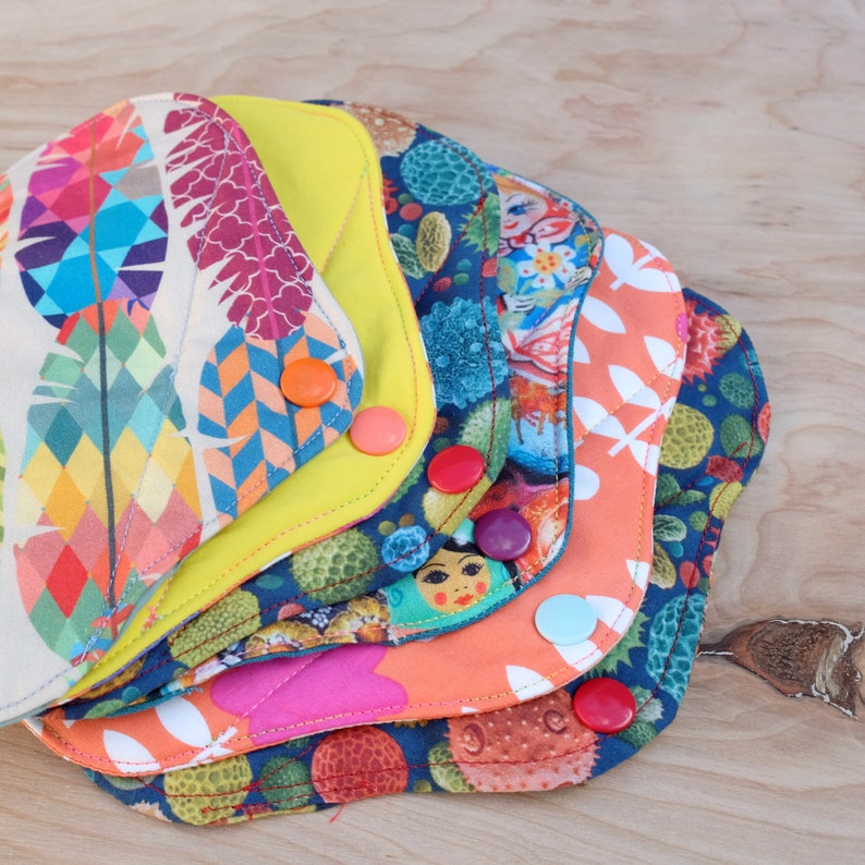 May include: A stack of six colourful fabric pads with different patterns, including feathers, flowers, and geometric designs. Each pad has a snap button closure.