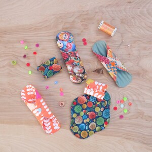 May include: Assortment of colourful reusable cloth menstrual pads with various patterns, including a floral design, a pattern of Russian nesting dolls, and a geometric pattern. The pads are folded and arranged on a wooden surface.