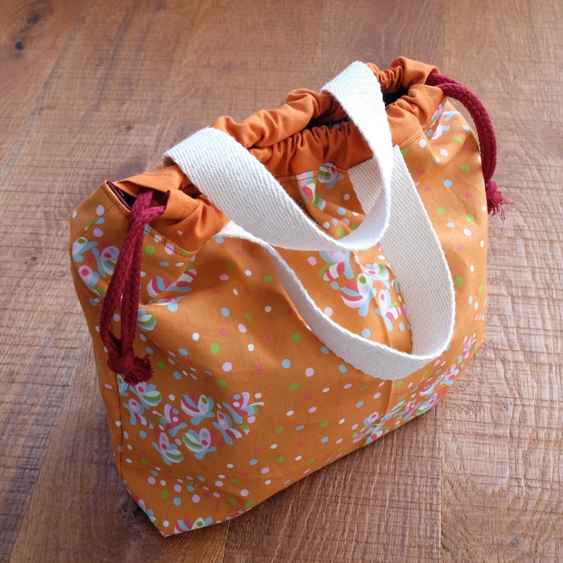 May include: A brown and orange drawstring bag with white straps and a colorful floral pattern. The bag has a red drawstring and is made of fabric.